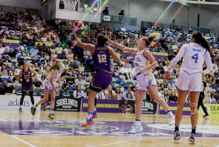 Women in singlets and shorts playing basketball on a court, with a large stadium crowd in the background.