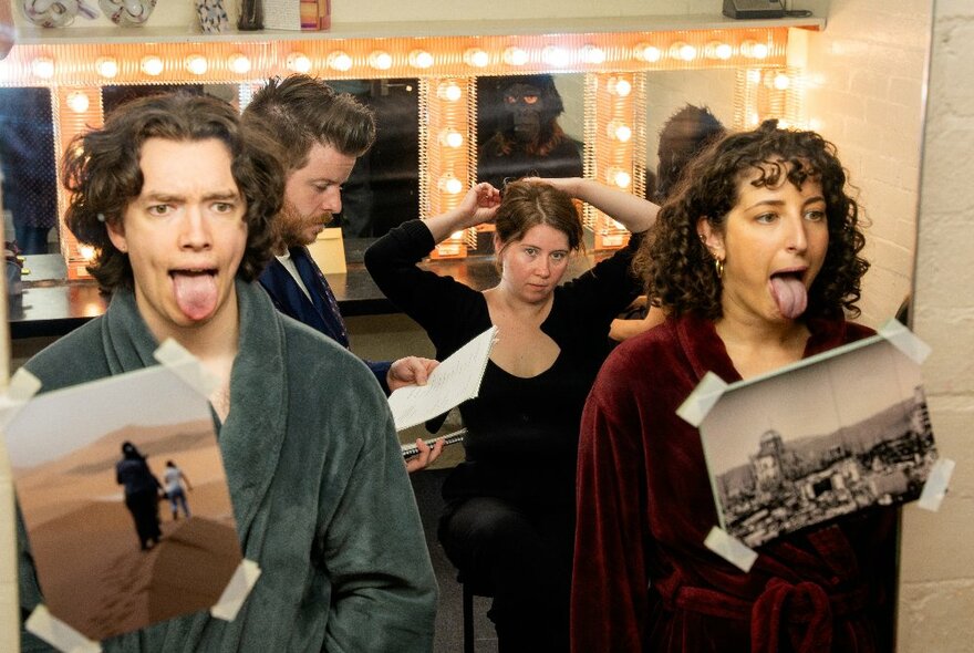 Four performers reflected in a mirror with photographs taped to it backstage, with two looking at themselves with tongues out.