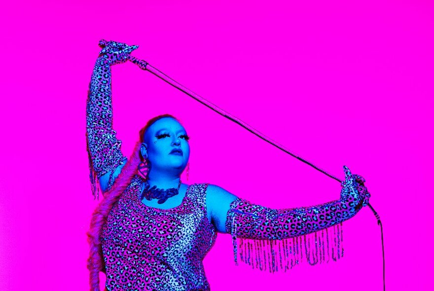 A woman wearing fringed long gloves and holds a whip above her head in front of a vivid pink background. 