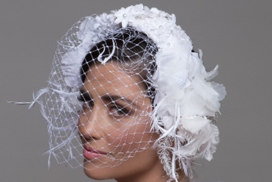 Model wearing a white feather fascinator with veil against a grey background.