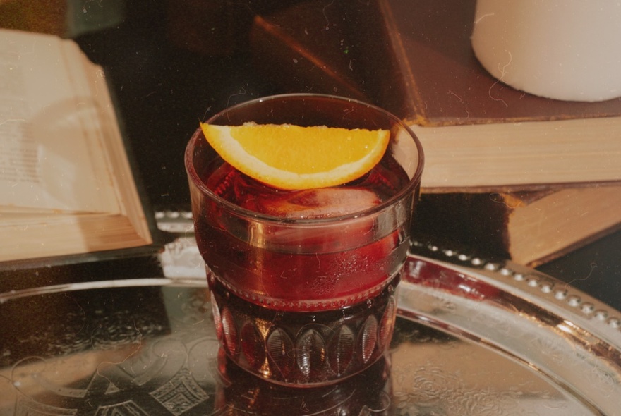 A cocktail in a short tumbler on a silver tray, garnished with a slice of lemon, with old books stacked in a pile next to it.