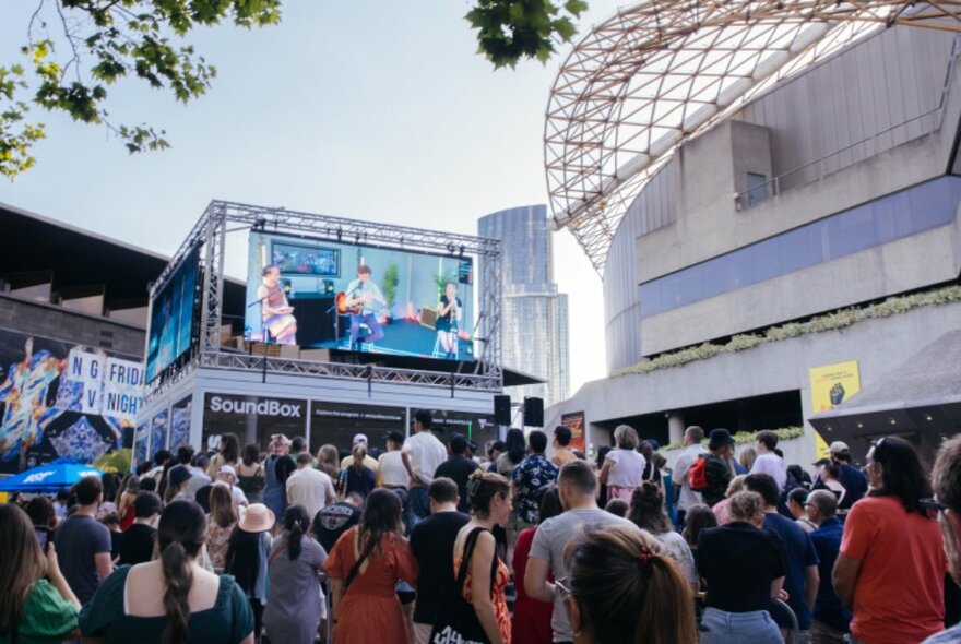 A crowd of people gathered in the forecourt of the  Arts Centre, two large screens displayed high above a cube-like structure, the Soundbox, in the background. 