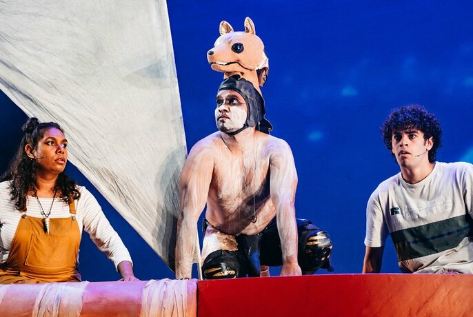 Three performers on a stage, in a prop boat with a sail, the central performer dressed as a dog.