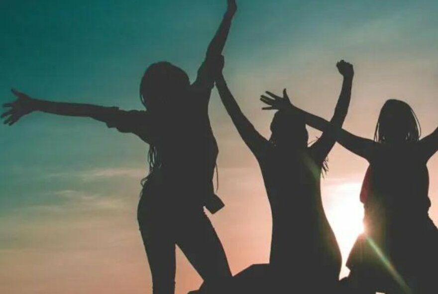 Three women standing with arms outstretched, silhouetted against a setting sun.