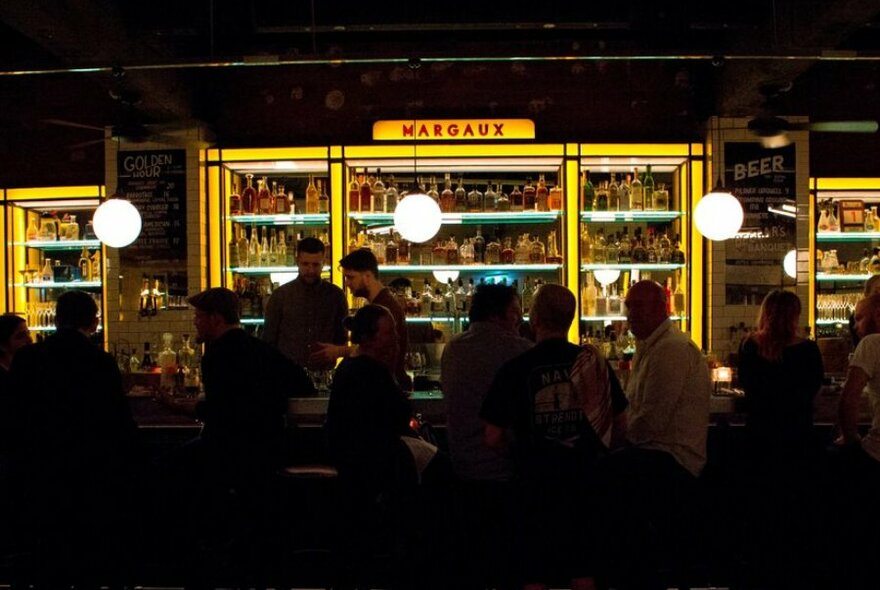
People sitting at a dark bar with neon yellow shelves filled with bottles of alcohol.