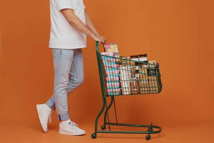 Person in pale jeans and a white t-shirt pushing a small supermarket style shopping trolley filled with goods, against an orange backdrop.