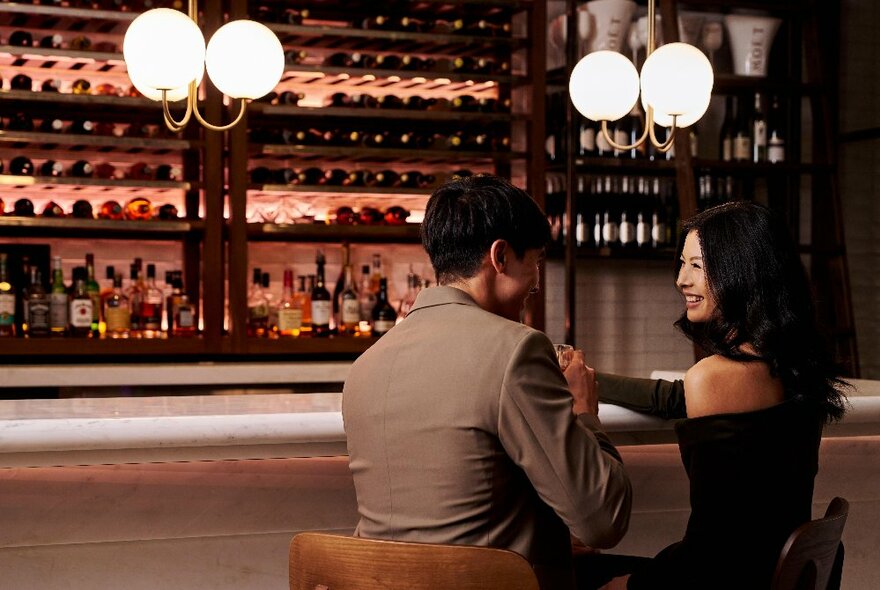 A couple seated at a bar counter in front of display cabinets filled with wine and liquor bottles, with round white glass pendant lighting.