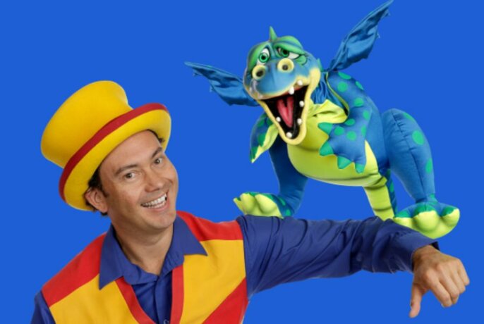 Comedian Tim Credible wearing a striped vest and yellow hat, with a dragon puppet on his arm, against a blue background.