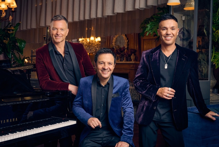 Singers Tim Campbell and Anthony Callea leaning near conductor John Foreman seated at a piano.