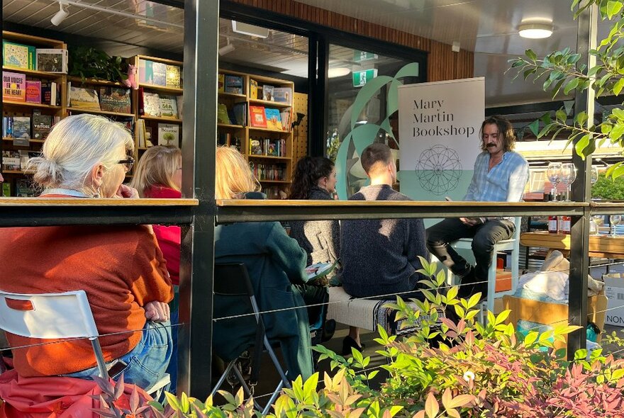 People seated on a verandah in front of a bookshop, listening to a person read from a book. 