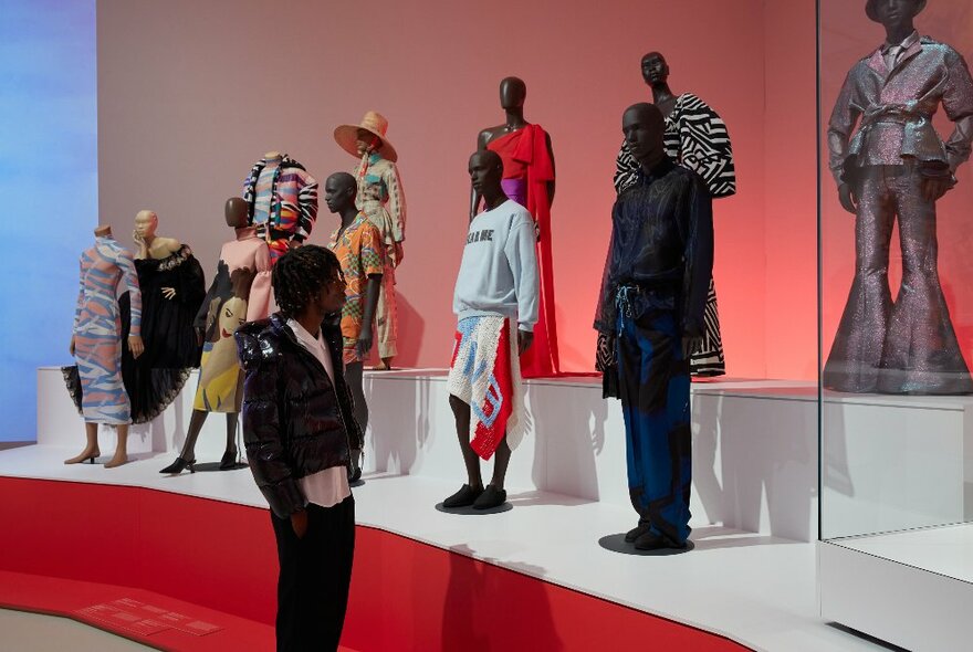 A gallery observer looking at a mannequins wearing different outfits displayed on a low platform in a gallery setting.
