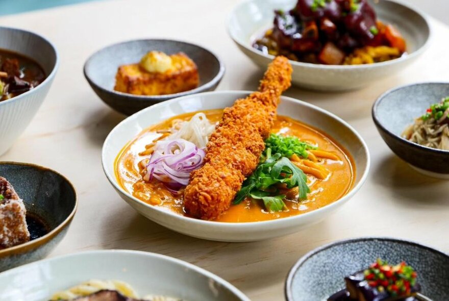 Crumbed item atop curry in bowl, surrounded by several other dishes.
