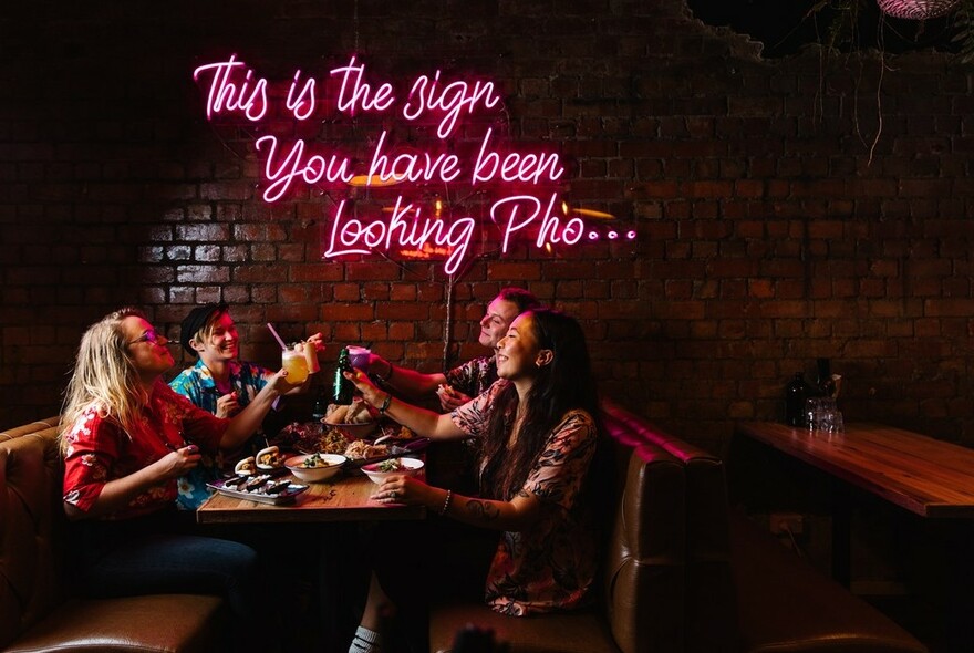 A group of four at a table clinking glasses in a toast underneath a pink neon sign. 