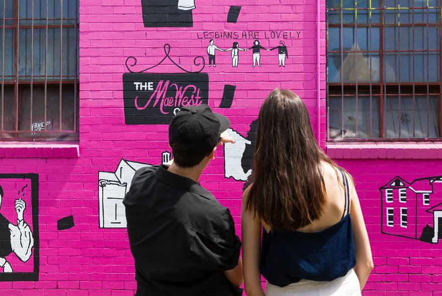 Two people looking at a wall painted hot pink with street art and signs on it.