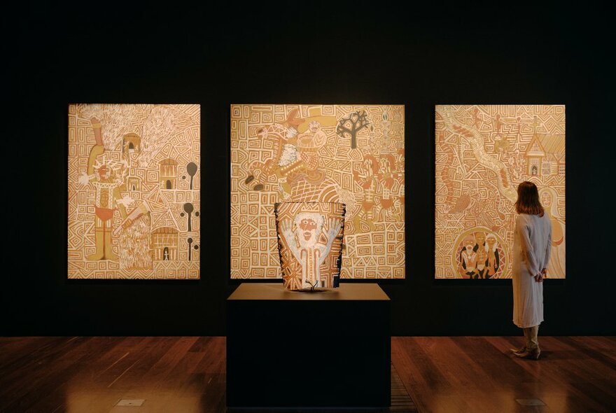 Three painted indigenous artworks side-by-side in a darkened gallery, with a bark painting on a pedestal in front; a woman contemplating one of the canvases.