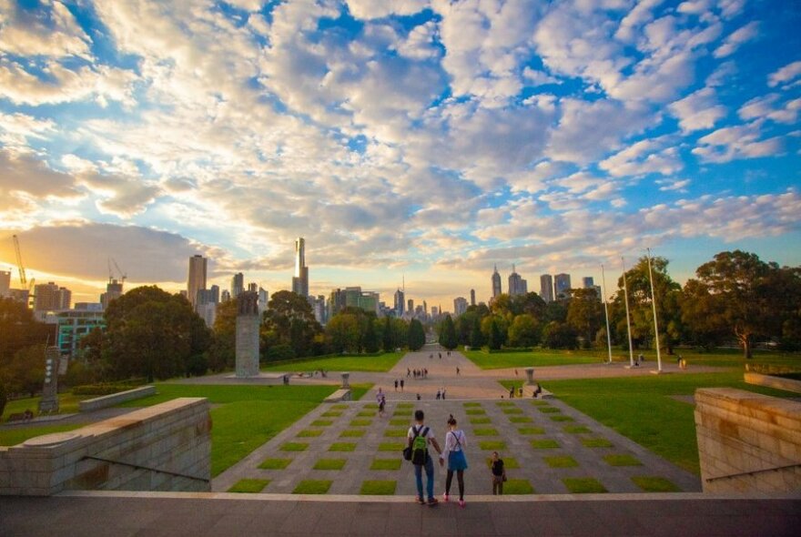 Two people standing on the edge of the Shrine, looking out over concrete and grass squares with Melbourne CBD buildings in the background.