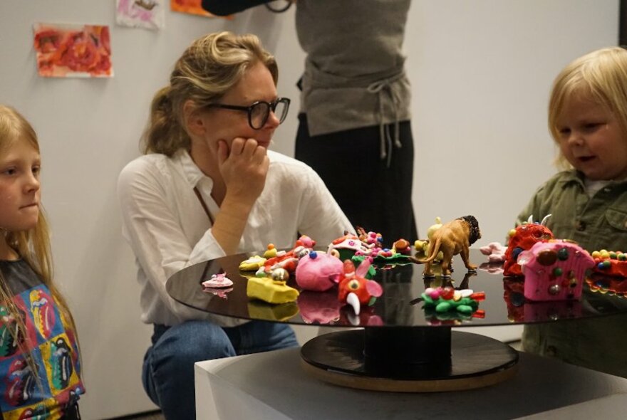 Adult crouched low watching two young children who are interacting with craft and models on a low table in a gallery space.