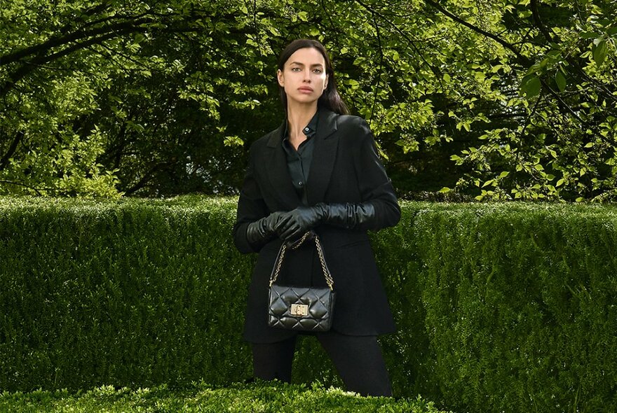 Model wearing a black coat and black elbow length leather gloves holding a small and elegant black handbag standing in front of a trimmed green hedge.