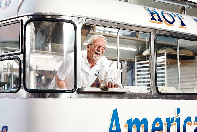 Smiling man with white hair and a white shirt leaning out of the service window of a food truck and holding a bag of donuts in his hand.