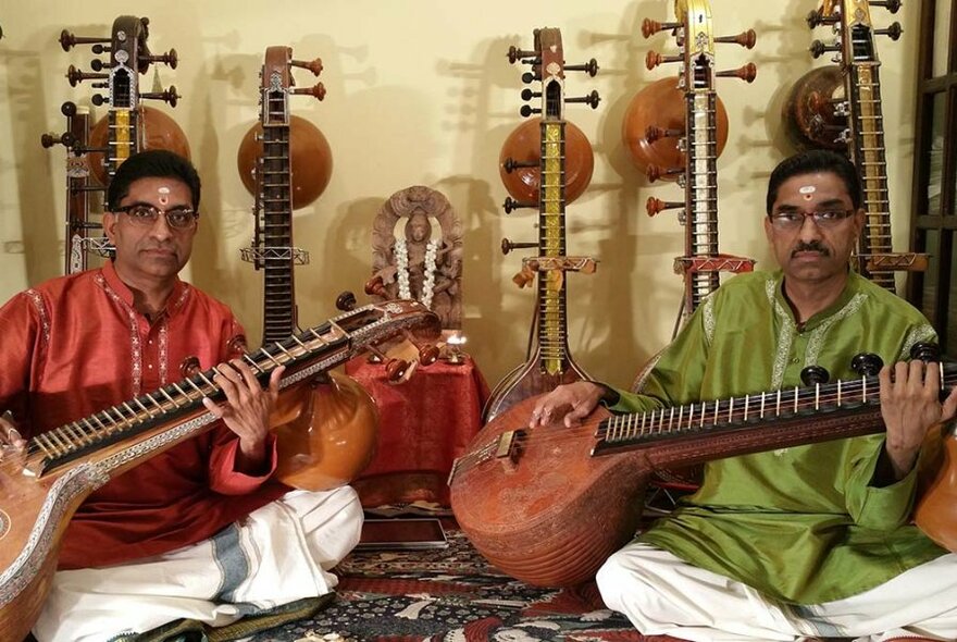 Two men sitting cross-legged on the floor playing traditional Indian veenas with more instruments leaning on the wall behind. 