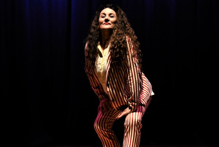 Grace Knight, woman with long, dark, wavy hair, wearing a white and red, thick pinstriped suit, striking a pose.