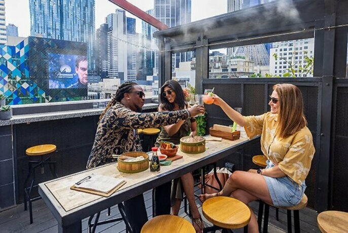 Three people sitting at a table in an outdoor courtyard of a rooftop bar, clinking their glass beverages together, with city buildings visible in the background.