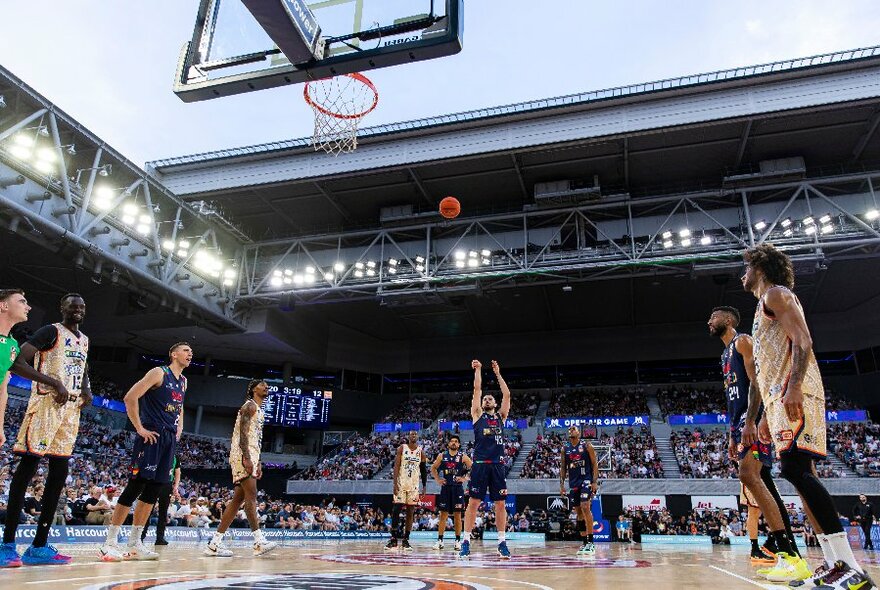 NBL basketball players on the court of an indoor stadium, one of them taking a free throw towards the hoop, with the roof open and the sky visible, and a seated audience watching the game.