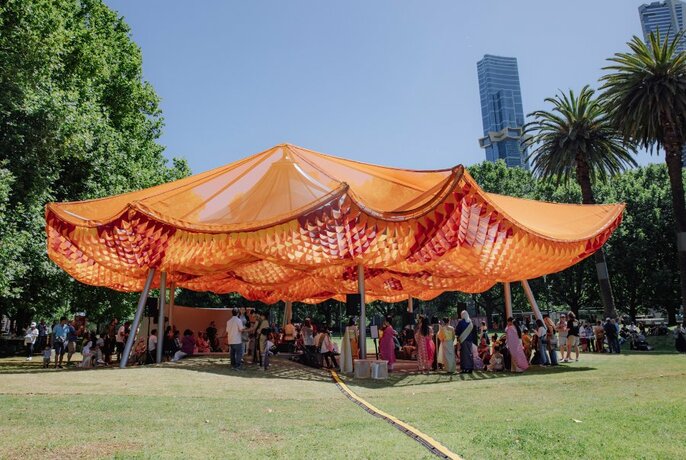 The MPavilion building, featuring a saffron-coloured sail design, in a park with people gathering around.