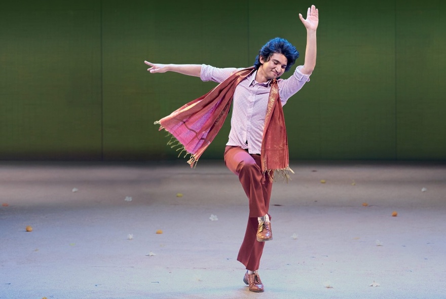 A dancer wearing pink scarf, shirt and trousers on stage, waving his arms.