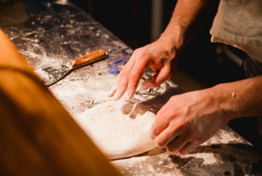 Hands flouring and kneading pizza dough on a wooden bench.