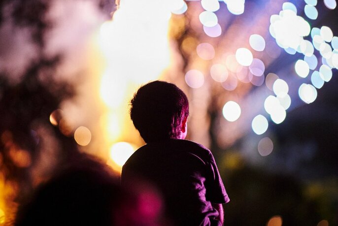 Silhouette view of a toddler from behind looking up at a night sky illuminated with fireworks.