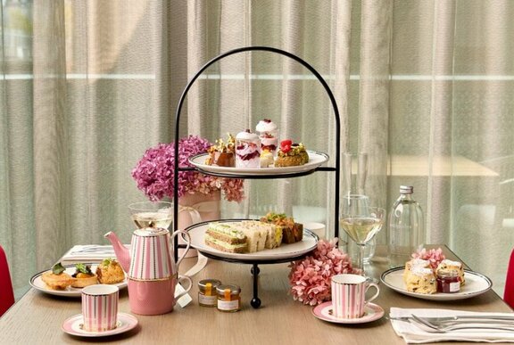 Table set with a high tea service, tiered plate of food, a vase of flowers, glasses of sparkling wine and tea and coffee.