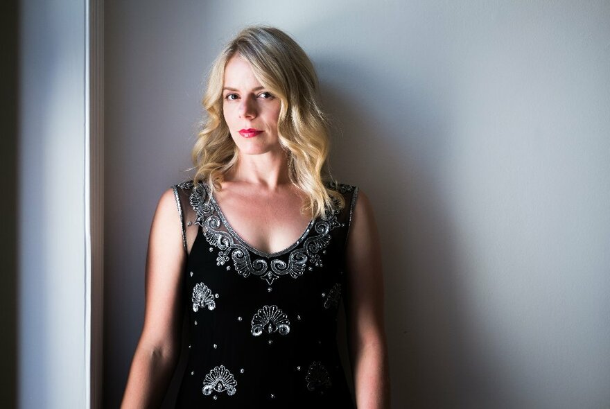 Women with shoulder length blonde hair, wearing a black sleeveless v-neck dress with decorative overstitching on it, posed against a light grey coloured wall.