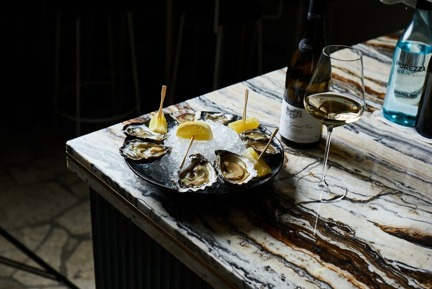 Oysters on a plate near the edge of a marble table with white wine in a glass and a bottle. 