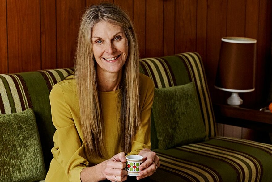 A smiling woman wearing a mustard coloured top, seated on a striped green velvet couch, drinking from a small espresso coffee cup.