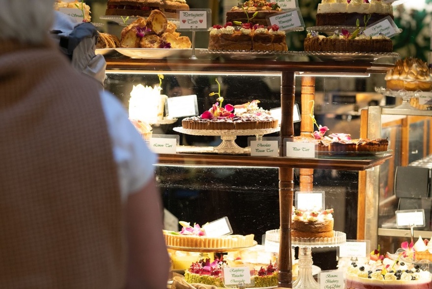 Rear view of a person looking through a glass window at a display of cakes and tarts on serving stands inside a cafe.