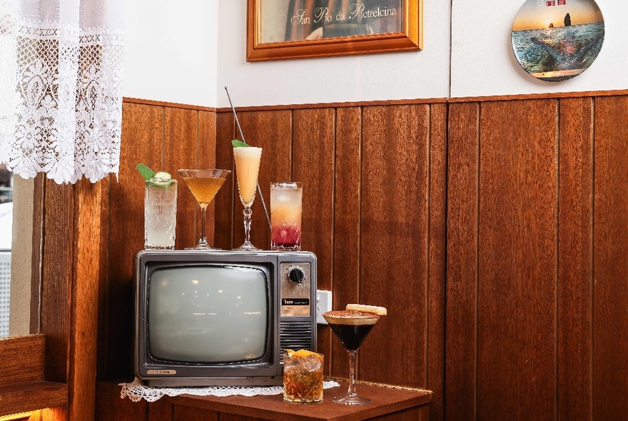 An old cube style TV on a wooden table in the corner of a wood paneled room, with a selection of six cocktails displayed around or on top of the TV.