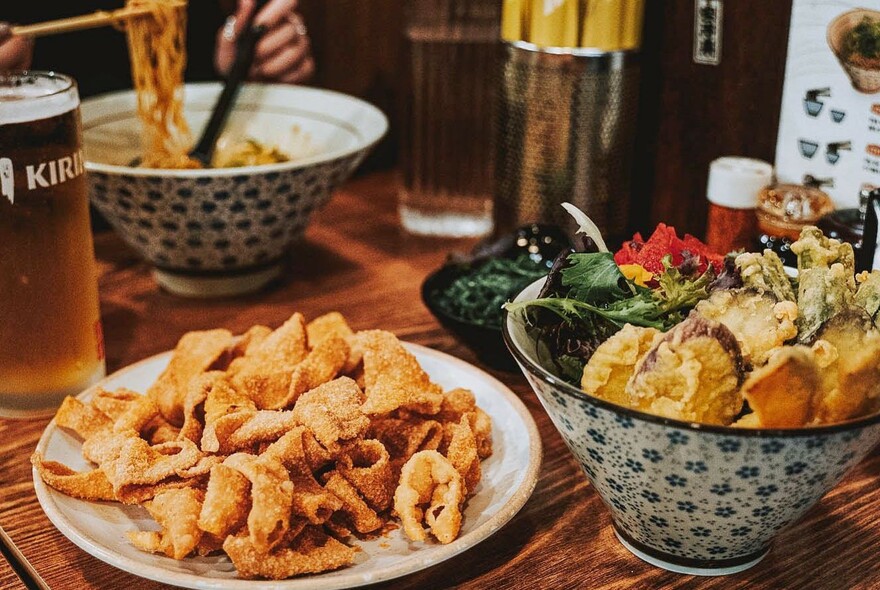 Wooden table with bowls of ramen and plate of fried food.