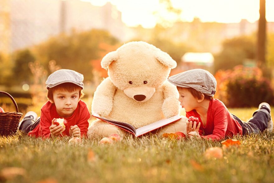 Two young children lying in the grass outdoors, wearing matching red tops and caps, both eating apples, with a large teddy bear holding a story book seated between them.
