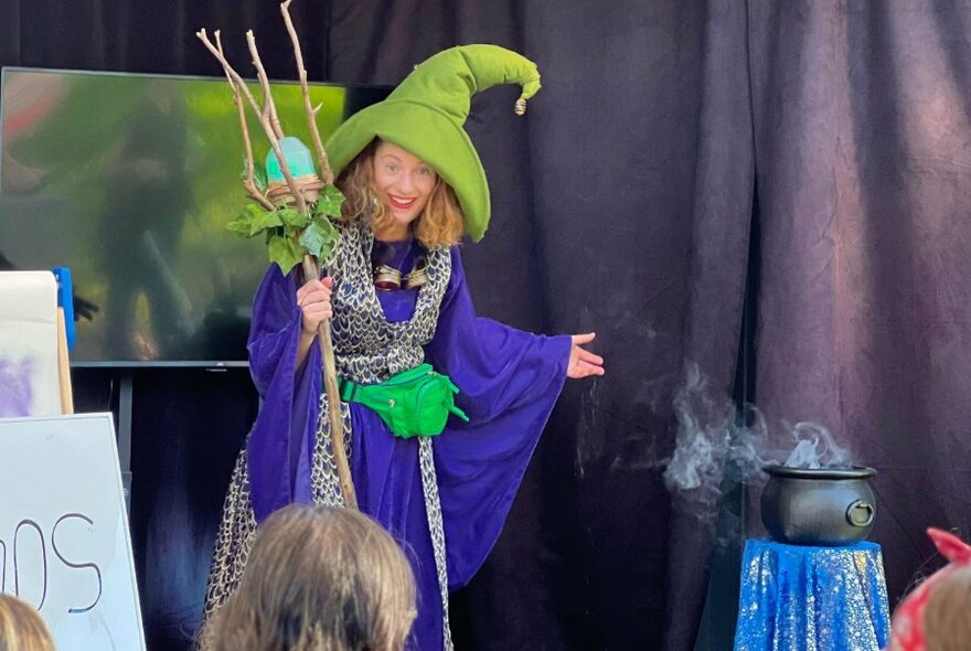 A wizard performer wearing a big green hat standing next to a smoking cauldron in front of an audience.