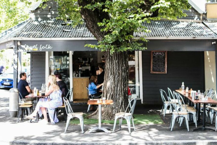 A small house cafe with a tree directly out the front and people sitting at tables in the dappled sunlight.