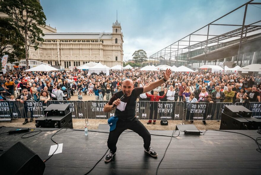 Performer on stage outside the Royal Exhibition Building, with crowds of people standing looking on under white umbrellas.