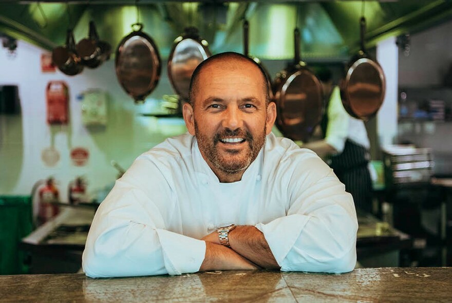 Smiling chef in a kitchen with copper pots hanging in the background.