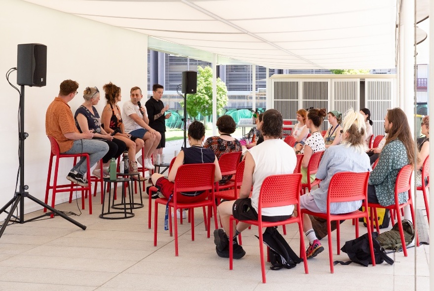 A panel discussion with five people seated on red stools in front of an audience seated on red chairs, under the flat roof structure of the MPavilion.