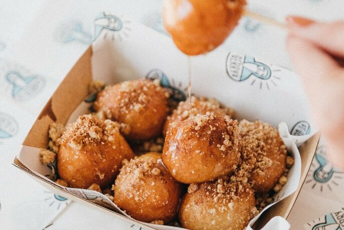 Takeaway box of syrupy small donuts garnished with chopped nuts.