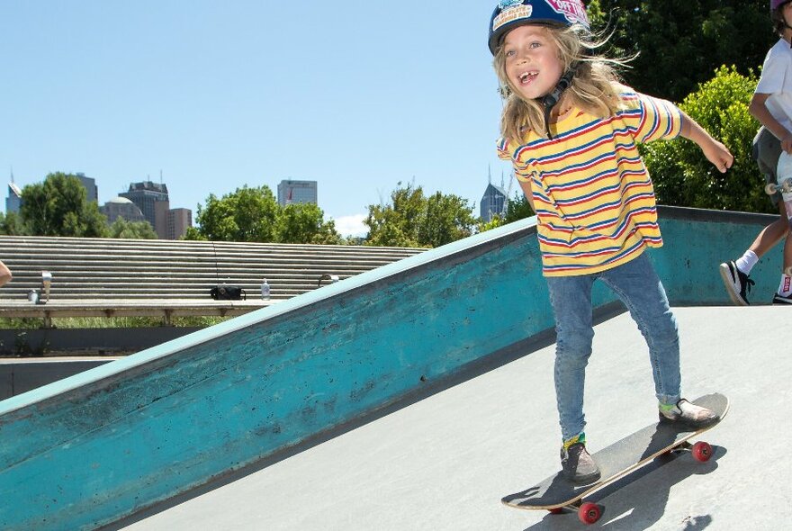 Young girl wearing a striped t-shirt, jeans and a safety helmet riding a skateboard down a ramp.