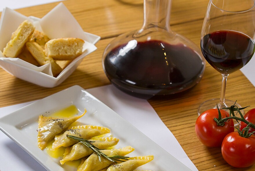White tray of pasta with decanter and glass of red wine, tomatoes and bread.