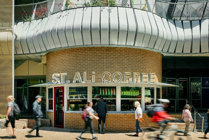 Exterior of the St ALi coffee kiosk at University of Melbourne showing people walking and cycling past a curved brick structure that has a takeaway window counter. 