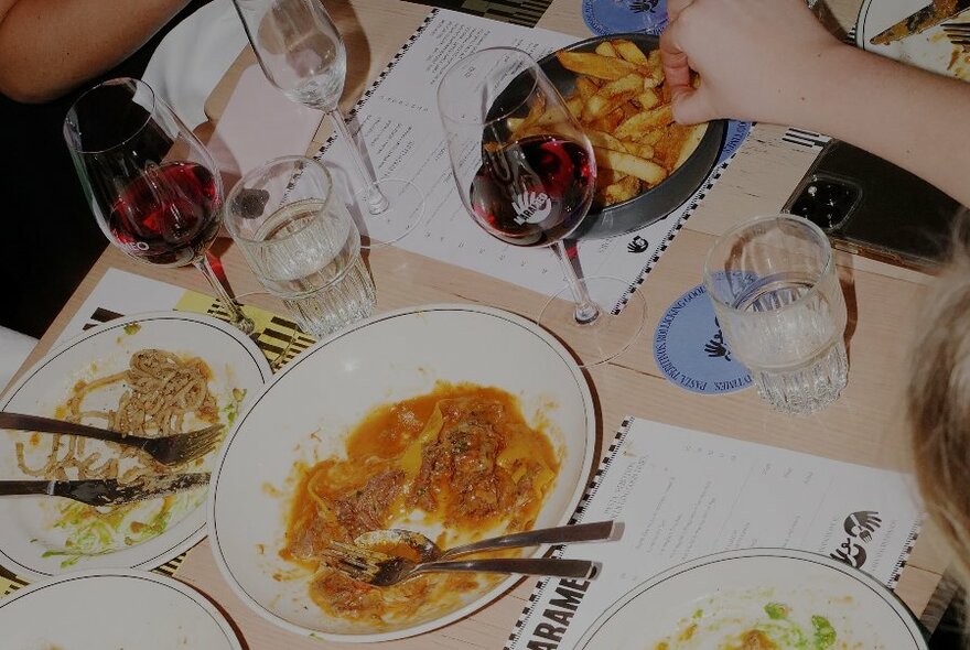 Looking down at a restaurant table of dishes that include spaghetti, meatball pasta, chips, and glasses of wine.
