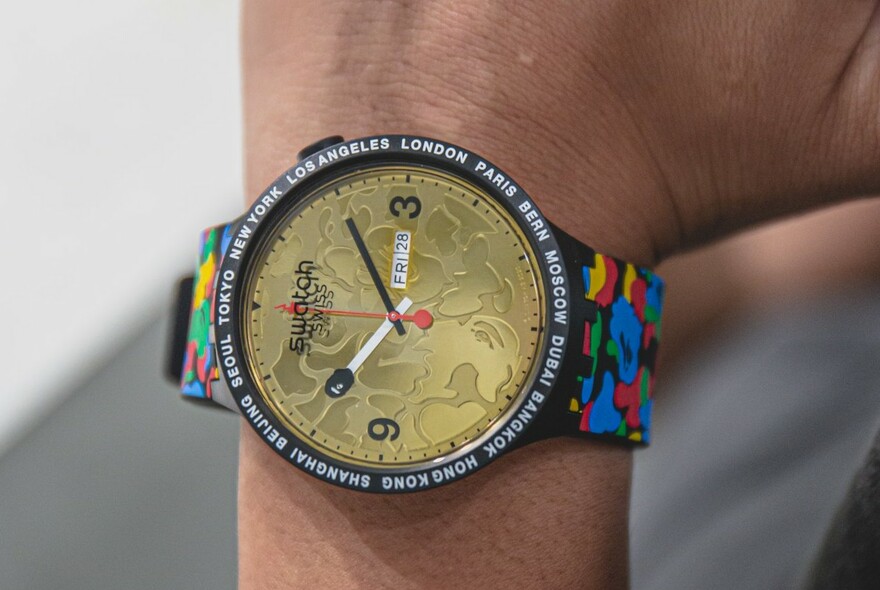 Wrist wearing a Swatch with a gold coin face and multicoloured strap.